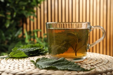 Photo of Glass cup of aromatic nettle tea and green leaves on wicker mat