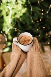 Photo of Woman holding cup of cocoa near Christmas tree indoors, closeup