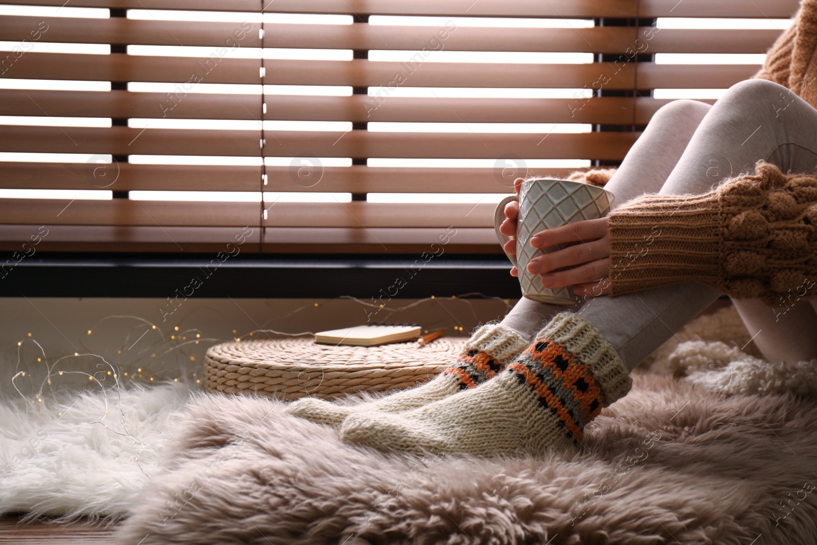 Photo of Woman with cup of hot drink wearing knitted socks on window sill indoors, closeup. Warm clothes