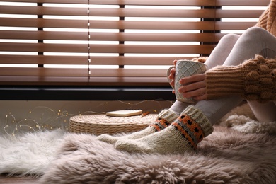 Woman with cup of hot drink wearing knitted socks on window sill indoors, closeup. Warm clothes