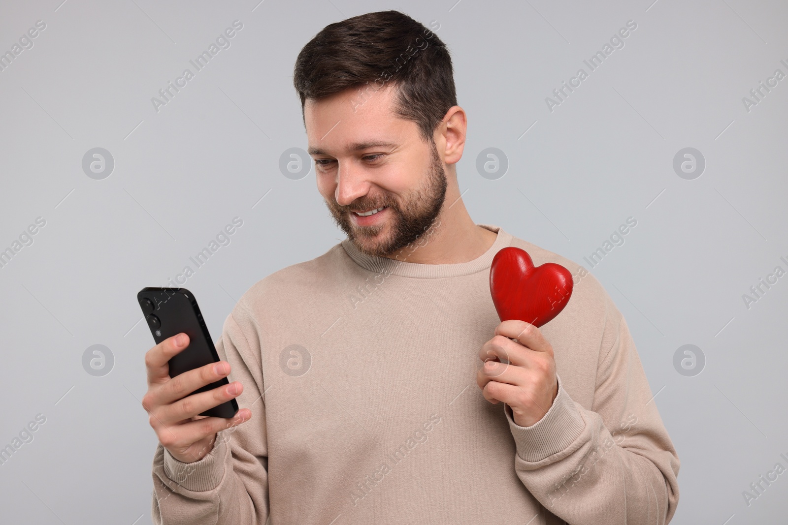 Photo of Happy man using smartphone and holding red heart on light grey background