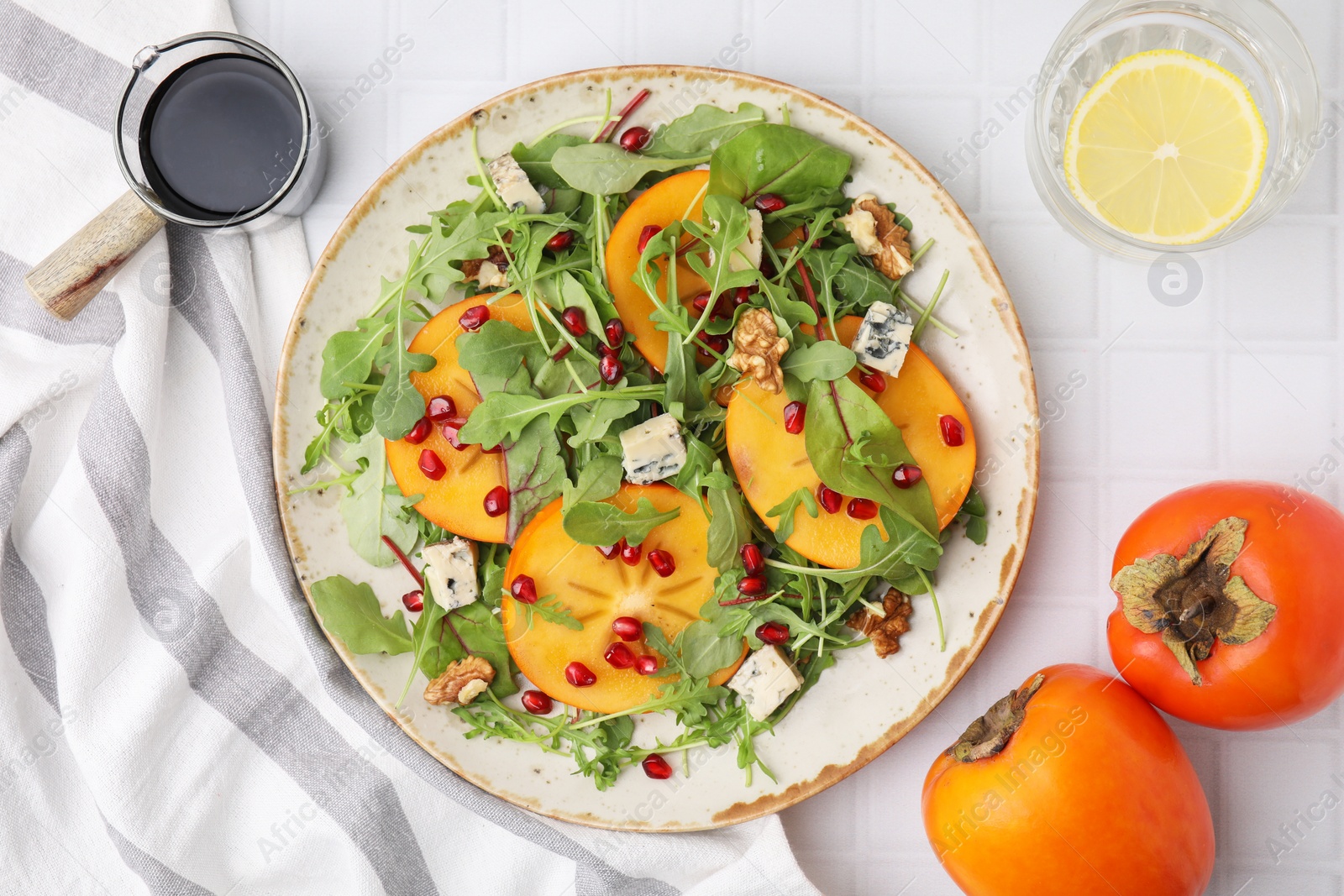 Photo of Tasty salad with persimmon, blue cheese, pomegranate and walnuts served on white tiled table, flat lay