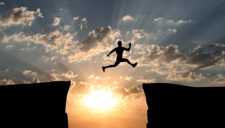 Image of Concept of reaching life and business goals. Silhouette of man jumping over chasm at sunrise