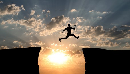 Concept of reaching life and business goals. Silhouette of man jumping over chasm at sunrise