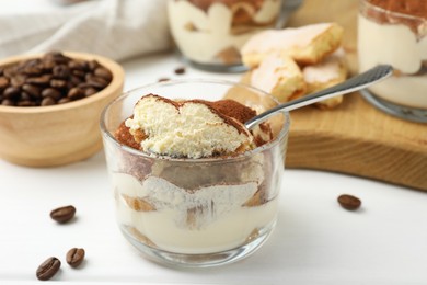 Photo of Delicious tiramisu in glass, spoon, coffee beans and cookies on white table, closeup