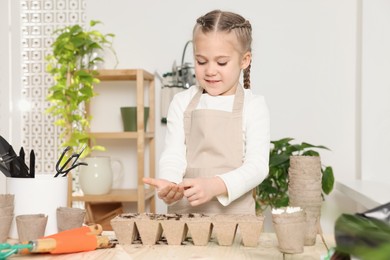 Photo of Little girl planting vegetable seeds into peat pots with soil at wooden table in room