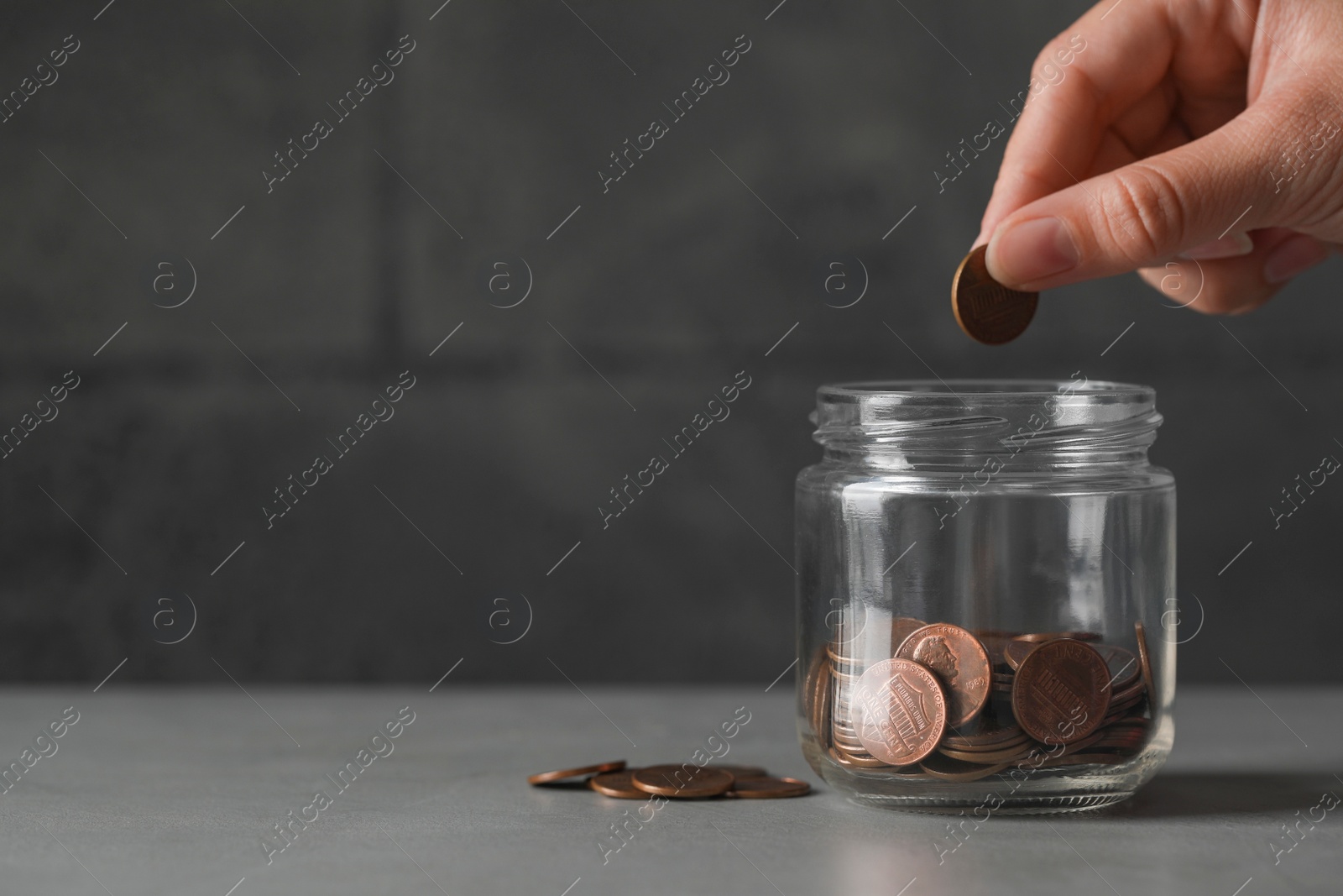 Photo of Woman putting coin in glass jar with money at grey table, closeup. Space for text