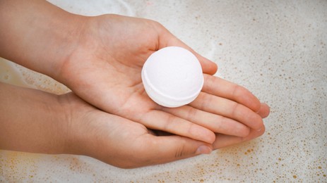 Woman holding bath bomb over water with foam, closeup