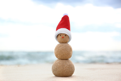 Snowman made of sand with Santa hat on beach near sea. Christmas vacation