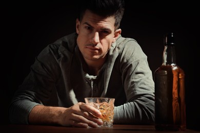 Photo of Addicted man with glass of alcoholic drink at wooden table against dark background