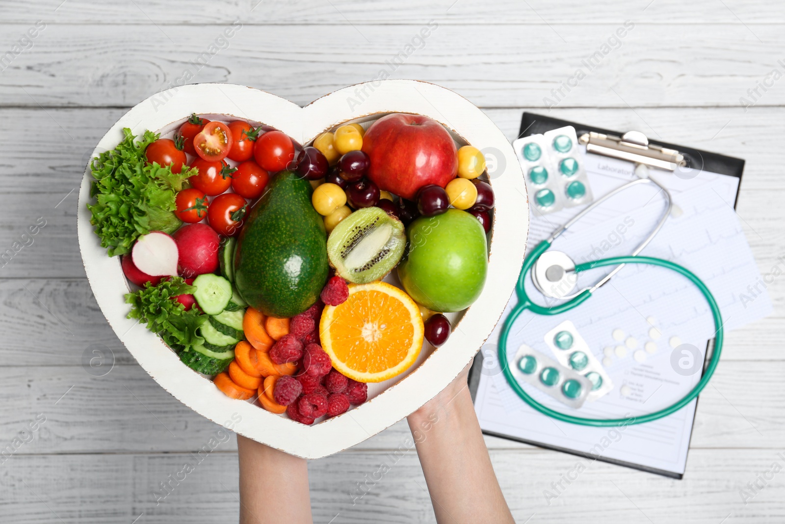 Photo of Female doctor holding plate with fresh fruits and vegetables over table, top view. Cardiac diet