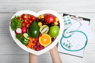 Photo of Female doctor holding plate with fresh fruits and vegetables over table, top view. Cardiac diet