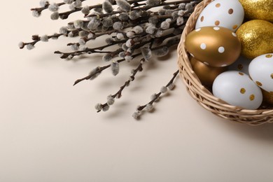 Photo of Wicker basket with decorated Easter eggs and willow branches on beige background, closeup. Space for text