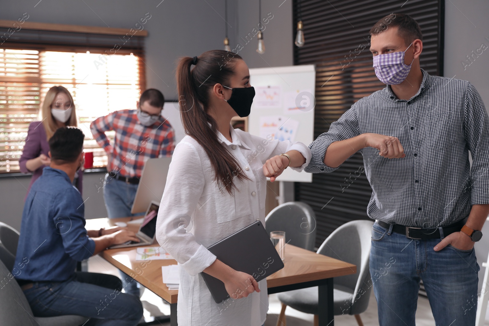 Photo of Coworkers with protective masks making elbow bump in office. Informal greeting during COVID-19 pandemic