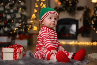 Photo of Cute little baby wearing Santa's elf clothes with Christmas gift on floor at home