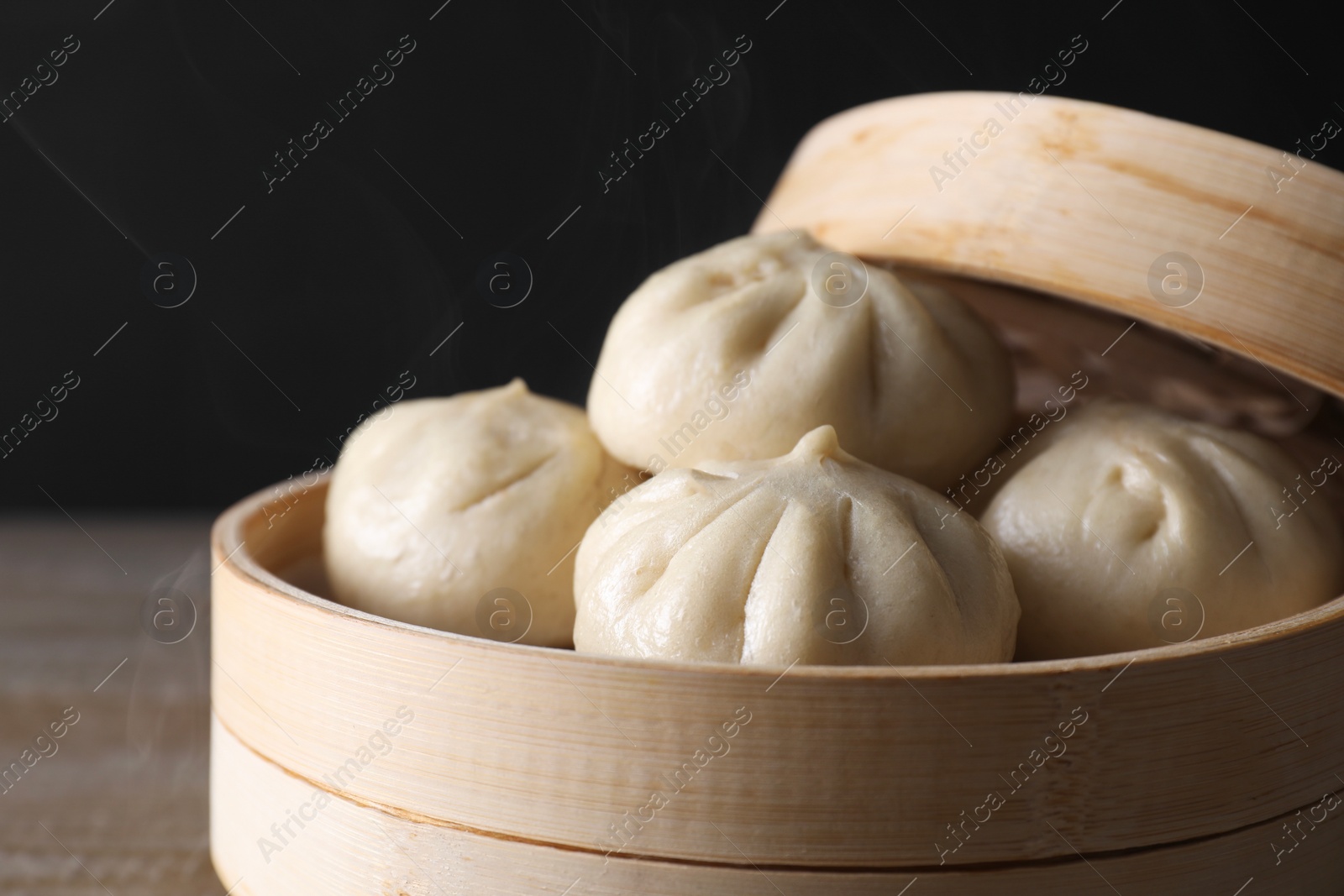 Photo of Delicious bao buns (baozi) in bamboo steamer on wooden table against black background, closeup