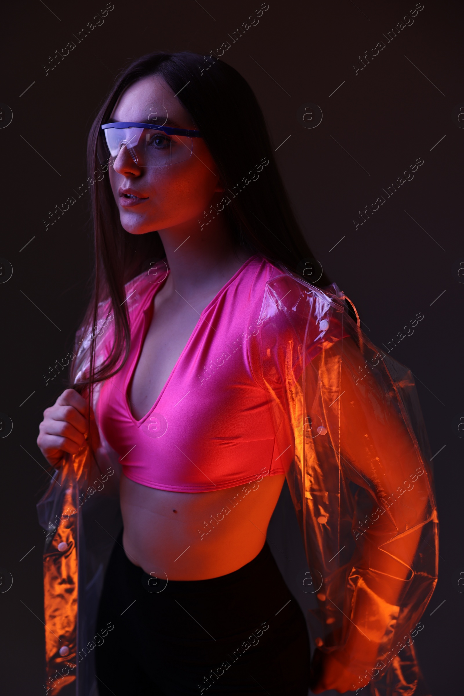 Photo of Fashionable portrait of beautiful woman wearing transparent coat and glasses on dark background in neon lights