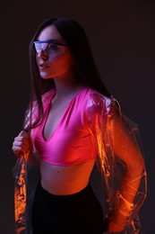 Fashionable portrait of beautiful woman wearing transparent coat and glasses on dark background in neon lights