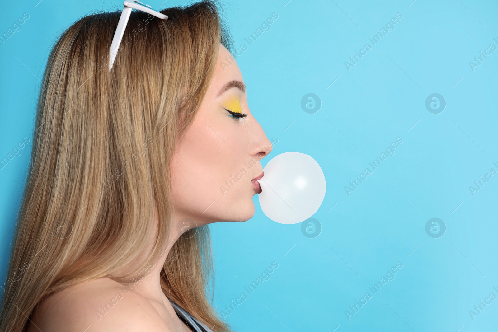 Photo of Fashionable young woman with bright makeup blowing bubblegum on light blue background, space for text