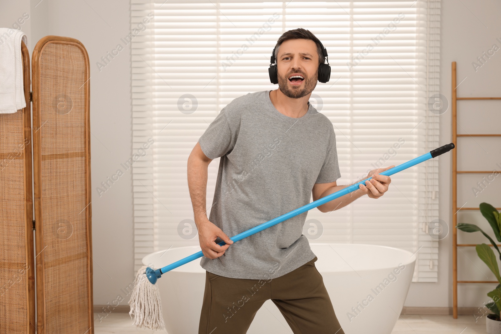 Photo of Enjoying cleaning. Happy man in headphones with mop listening to music while tidying up in bathroom