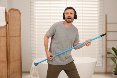 Photo of Enjoying cleaning. Happy man in headphones with mop listening to music while tidying up in bathroom