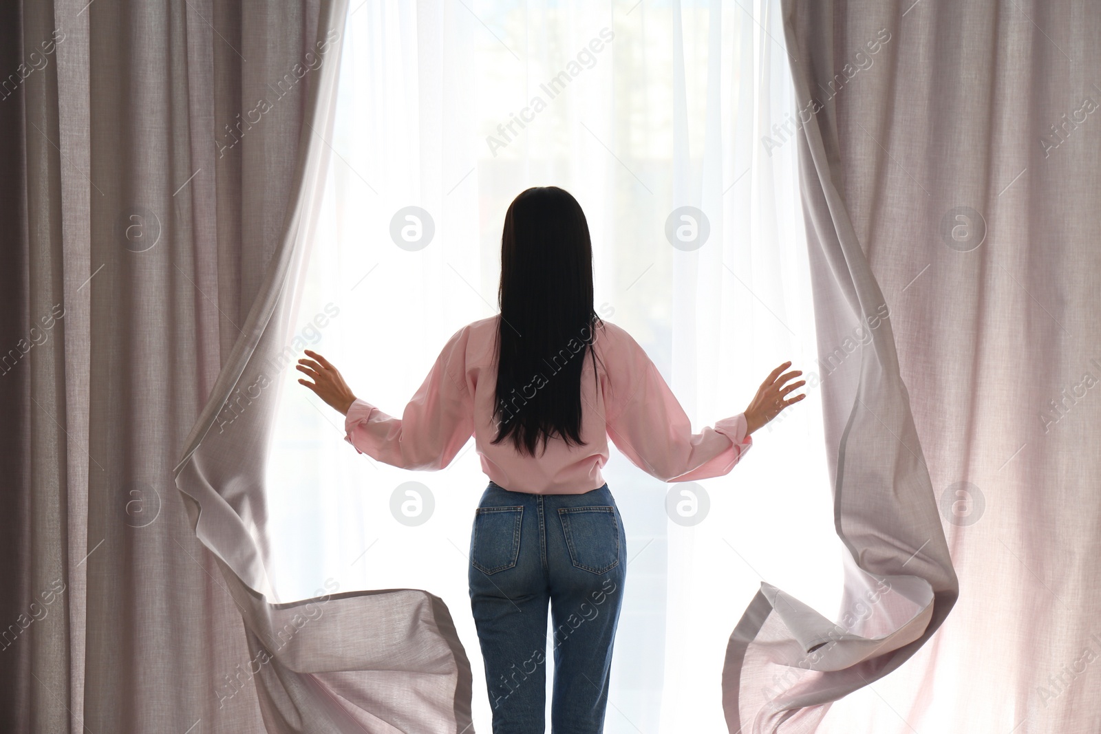 Photo of Woman opening window curtains at home in morning, back view