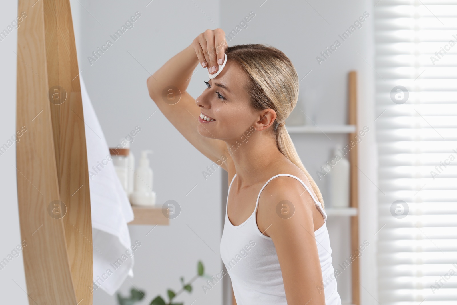 Photo of Smiling woman removing makeup with cotton pad in front of mirror in bathroom