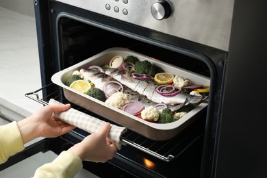 Photo of Woman putting baking tray with sea bass fish and vegetables into oven, closeup