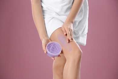 Young woman applying body scrub on leg against color background