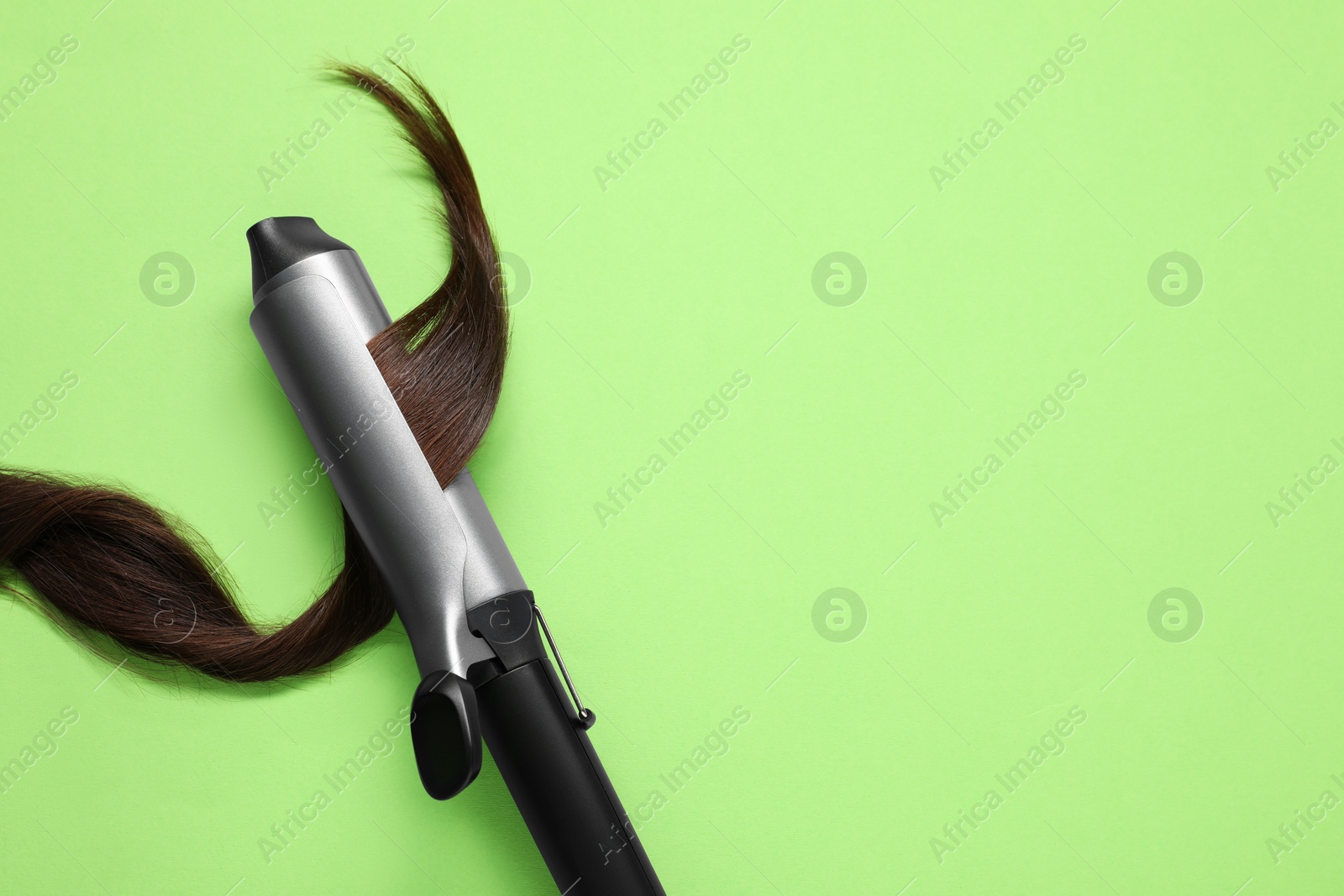 Photo of Curling iron with brown hair lock on green background, top view. Space for text