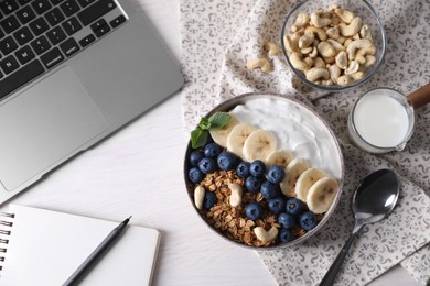 Delicious granola in bowl, stationery and laptop on white wooden table, flat lay