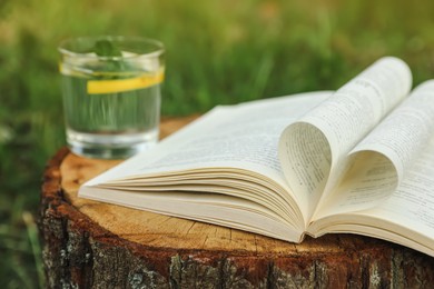Photo of Open book near glass of water with mint and lemon on tree stump outdoors, closeup