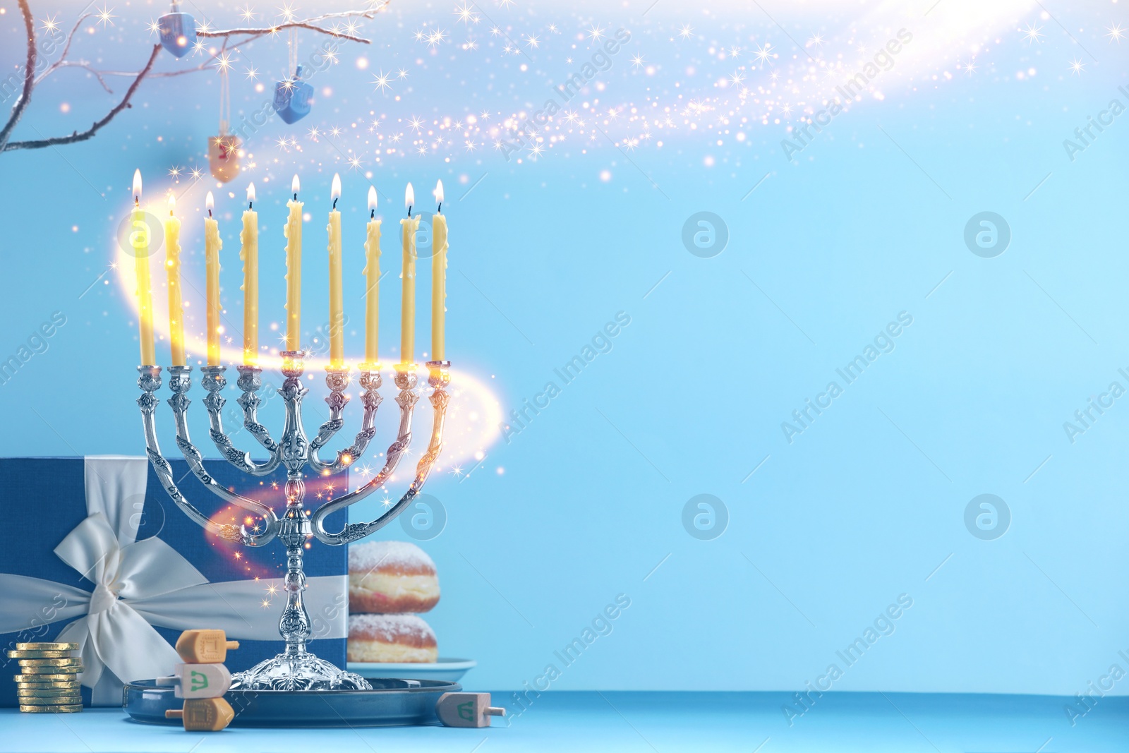 Image of Hanukkah celebration. Menorah with burning candles, dreidels, gift box and donuts on table against light blue background, space for text