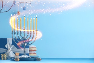 Hanukkah celebration. Menorah with burning candles, dreidels, gift box and donuts on table against light blue background, space for text