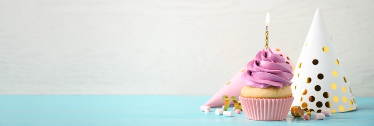 Image of Delicious birthday cupcake with burning candle on light blue wooden table, space for text. Banner design