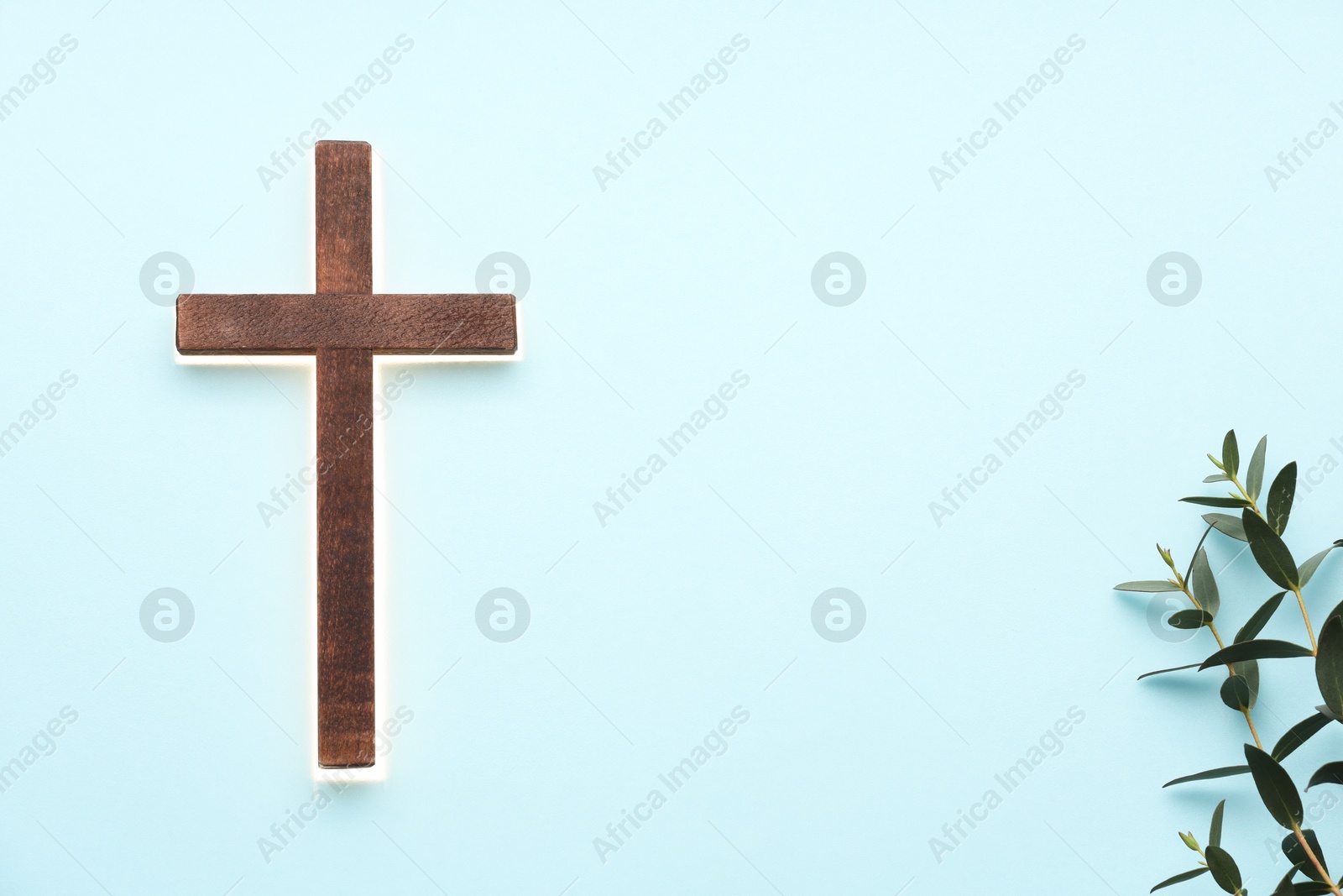 Photo of Shining cross and eucalyptus branches on light blue background, space for text. Religion of Christianity