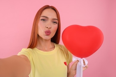 Photo of Beautiful woman with red heart shaped balloon taking selfie and sending air kiss on pink background