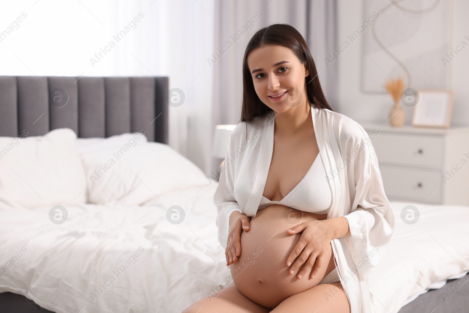 Photo of Beautiful pregnant woman in stylish comfortable underwear and robe on bed at home, space for text