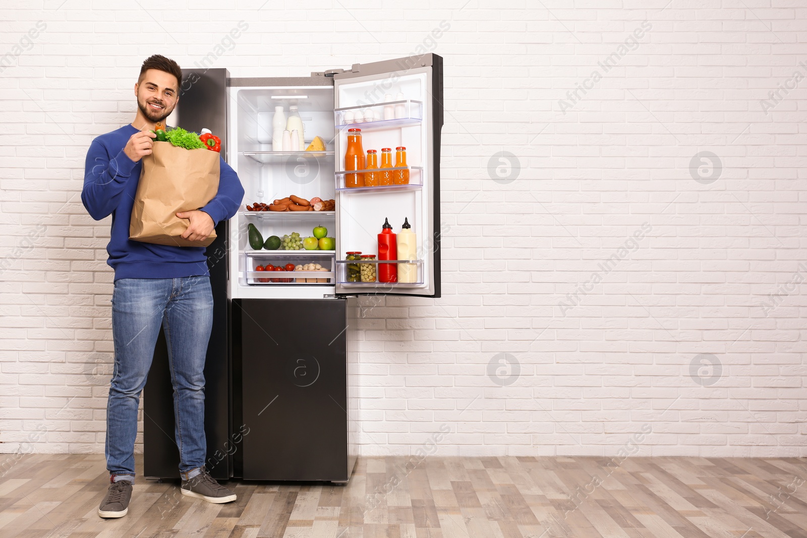 Photo of Young man with paper bag full of products near open refrigerator indoors, space for text