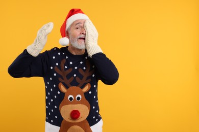 Photo of Senior man in Christmas sweater, Santa hat and knitted mittens on orange background. Space for text