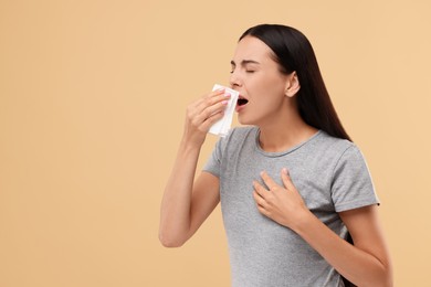 Photo of Suffering from allergy. Young woman blowing her nose in tissue on beige background. Space for text
