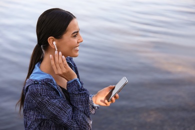 Photo of Young sportswoman with wireless earphones and smartphone near river