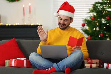 Photo of Celebrating Christmas online with exchanged by mail presents. Happy man in Santa hat with greeting card during video call on laptop at home