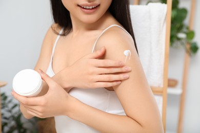 Photo of Young woman applying body cream on shoulder in room, closeup