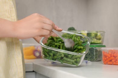 Photo of Woman taking green beans with spoon from glass container at white marble table in kitchen, closeup. Food storage