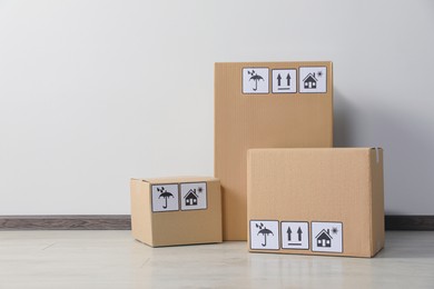 Photo of Many closed cardboard boxes with packaging symbols on floor near white wall. Delivery service