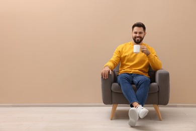 Photo of Handsome man with cup of drink sitting in armchair near beige wall indoors, space for text