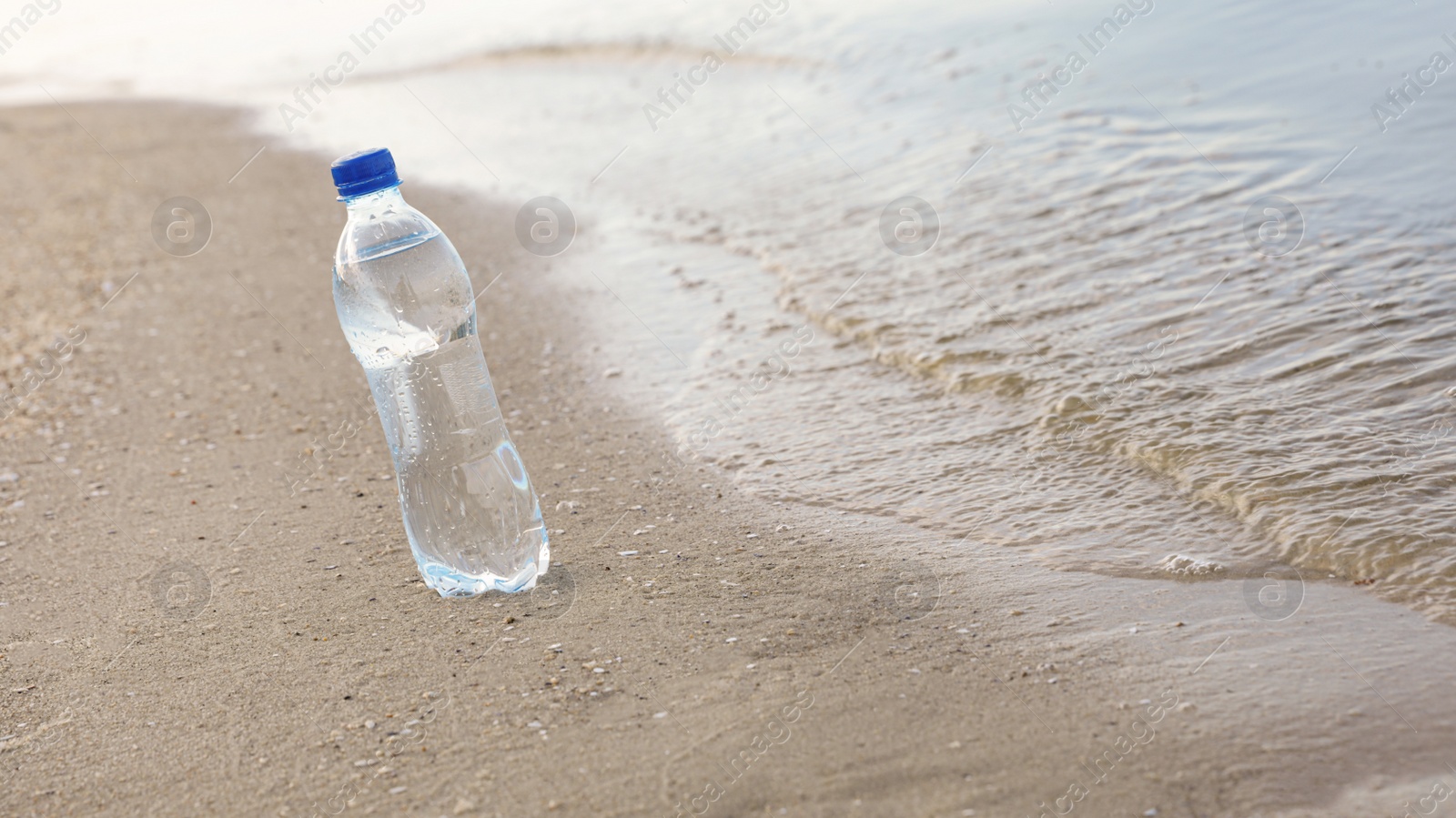 Photo of Plastic bottle with refreshing drink on sandy beach near sea, space for text. Hot summer day