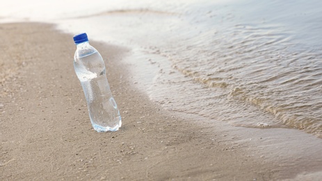 Plastic bottle with refreshing drink on sandy beach near sea, space for text. Hot summer day
