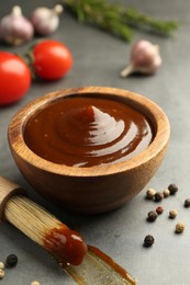 Tasty barbeque sauce in bowl, brush and peppercorns on grey table, closeup